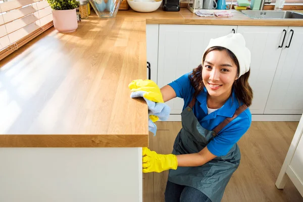 Professional Asian woman cleaning service wearing yellow rubber gloves, using a rag to wipe with spraying liquid detergent on the wooden top kitchen counter at home. Housekeeping cleanup, cleaner.