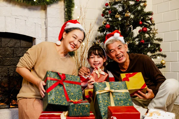 Happy and cheerful Asian family gathering, wearing Santa hats talking and smiling. Exchanging gifts, grandparents and grandson daughter, sitting at the fireplace with decorated Christmas tree.