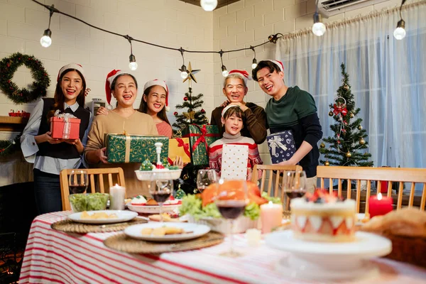 Happy and cheerful Asian family gathering, wearing Santa hats talking and smiling. Exchanging gifts, grandparents, grandchildren, daughters, at Christmas dinner table with decorated Christmas tree.