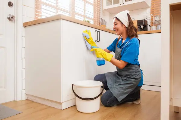 Profesyonel Asyalı kadın temizlik servisi sarı lastik eldiven giyiyor, evdeki ahşap mutfak tezgahına sıvı deterjan sıkmak için bez kullanıyor. Temizlik, temizlik..