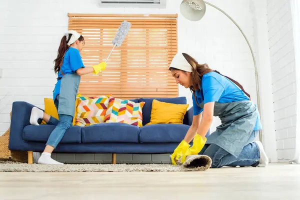 Two Asian young professional cleaning service women worker team working in the house. Girl housekeeper rolled keeping the carpet before cleaning wooden floor with another one cleaning the sofa pillow.