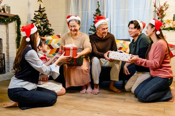 Happy and cheerful Asian family gathering, wearing Santa hats talking and smiling. Exchanging gifts, grandparents, grandchildren, daughters, sitting at the fireplace with decorated Christmas tree.