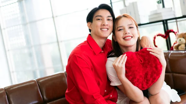A young happy Asian couple looking at the camera, sitting, embracing and smiling on Valentine\'s Day at home. Asian woman enjoys her anniversary date with her boyfriend. Valentine\'s Day celebration.