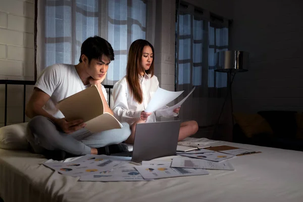 Young Asian couple work late and sit with a laptop and more data documents and presentations on the bed in the bedroom at night. Men and women working late concept. Image with copy space