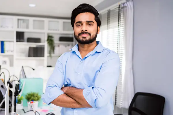 Successful Indian businessman entrepreneur in casual smiling and looking at the camera while standing with his arms crossed. Happy businessman standing in the office of a modern corporate workplace.