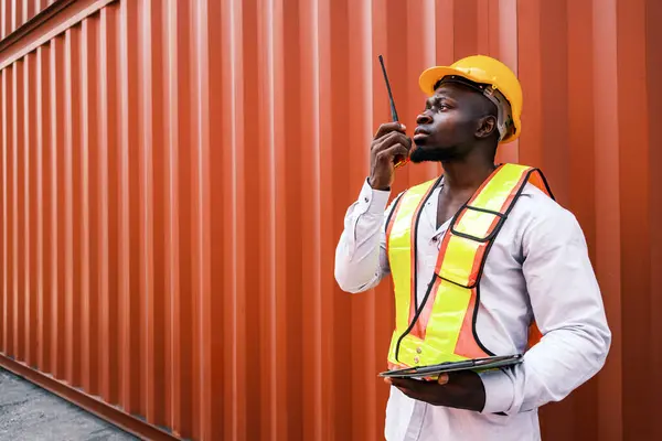 Portrait of an African man engineer or industrial worker in white hard hat, high-visibility Vest, talking on walkie-talkie radio at container yard. Inspector or safety supervisor in container terminal