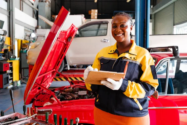 Portrait of an African mechanic woman in yellow and blue uniforms standing in the garage. A woman smiling while holding a maintenance checklist. Automobile repairing service. Vehicle maintenance.