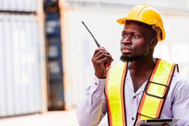 Afrikalı bir mühendisin portresi ya da sanayi işçisi, çelik yelek giymiş, konteynır deposunda telsizle konuşan. Konteyner terminalinde müfettiş ya da güvenlik danışmanı