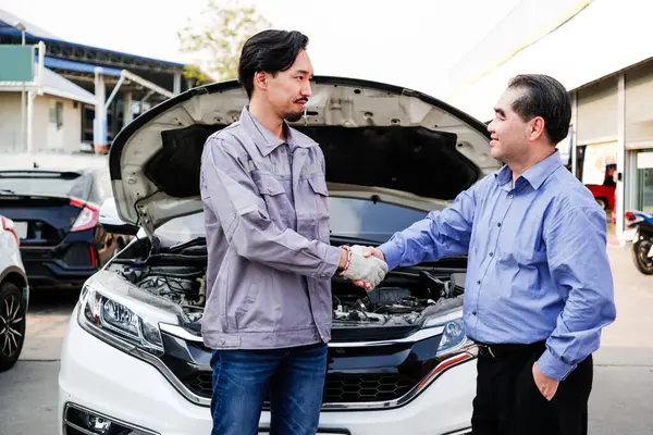 stock image Japanese mechanic car shaking hands with an Asian customer with an opened radiator hood car. Focus on a mechanic and a customer. Auto car repair service center. Professional service.