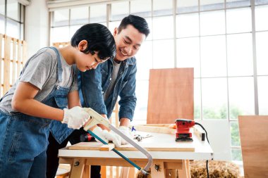 Asian father and son work as a woodworker or carpenter, Father teaches his son to saw a wooden plank with hack saw carefully together with teamwork. Craftsman carpentry working at home workshop studio