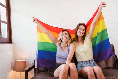 Kafkasyalı mutlu lezbiyen çiftin portreleri bir kanepede oturup kucaklaşıyorlar ve oturma odasında Gökkuşağı bayraklarını asıyorlar. LGBTQ dostluğu ya da çift ilişkileri birlikte kaliteli zaman geçirirler..