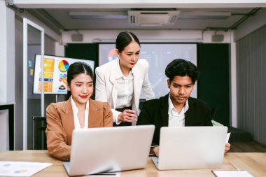 Neşeli ve kendine güvenen Asyalı bir iş kadını ofis arkadaşlarının arkasında duruyor ve bir monitör ve projektör ekranından bar çizelgeleri sunuyor. Toplantıda Asyalı işkadını lider rolü
