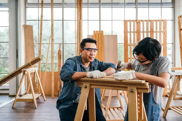 Happy Asian father and son work as a woodworker and carpenter. Father teaching his son to hammer nails on a wooden plank carefully together. carpentry working at a home workshop studio. Small business
