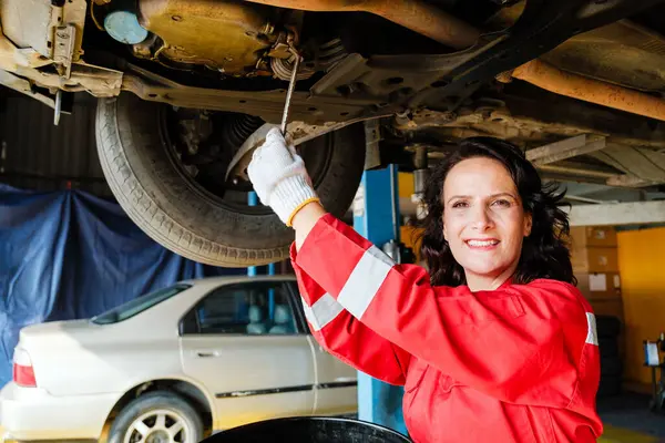 Garajı teftiş etmek için arabanın altında duran kırmızı üniformalı beyaz kadın bir tamircinin portresi. İngiliz anahtarını tutarken gülümseyen bir kadın. Araba tamir servisi. Araç bakımı