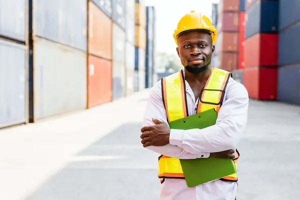 African man industrial worker standing, arm crossed works at a container yard. Shipping business management and international goods import-export. Logistic operation concept. Diversity labor workers