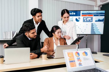 Neşeli ve kendine güvenen Asyalı bir iş kadını ofis arkadaşlarının arkasında duruyor ve bir monitör ve projektör ekranından bar çizelgeleri sunuyor. Toplantıda Asyalı işkadını lider rolü