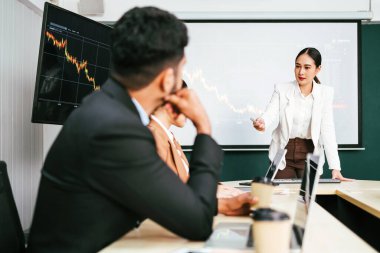 A cheerful and confident Asian businesswoman stands, present bar charts data from projector screen to her office colleagues. Asian business women leader role at the meeting.