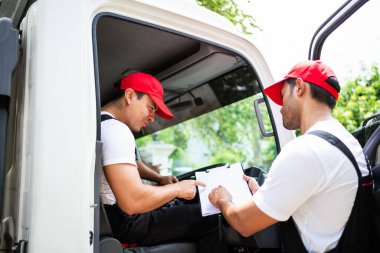 Kırmızı şapkalı asistanıyla birlikte mutlu bir profesyonel kamyon şoförü. Güler yüzlü ve teslimat yapılacaklar listesi ve yeri olan paket hakkında konuşuyor. Ev taşımacılığı ve teslimat konsepti için bir kamyon şoförü.