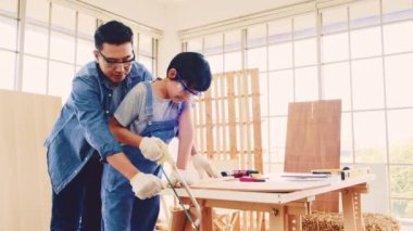 Asian father and son work as a woodworker or carpenter, Father teaches his son to saw a wooden plank with hack saw carefully together with teamwork. Craftsman carpentry working at home workshop studio