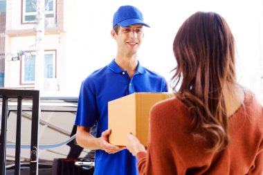 Mavi üniformalı teslimatçı. Kapüşonlu tişört giyen, kurye olarak çalışan bir erkek çalışanın elinde boş bir karton kutu var. Genç kurye eşikte paketler tutuyor. Teslimat hizmeti kavramı