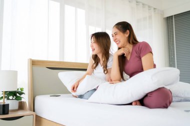 İki güzel Asyalı kadın, rahat bir yatak odasında beyaz bir yastıkla oturup eğlenerek gülüp gülüyorlar. Kız arkadaşlığı..