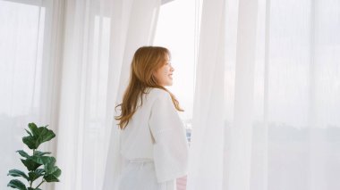 Bornoz giyen kadın yatak odasındaki perdeleri açar ve dışarıyı gezer. Hoş geldiniz yeni güneşli gün, mutlu olumlu genç bayan uyanıyor ve pencereden bakıyor evde ya da otelde iyimser hissediyor.