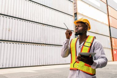 Beyaz kasketli, yüksek görüşlü yelek giyen, konteynır avlusunda telsizle konuşan Afrikalı bir mühendisin ya da sanayi işçisinin portresi. Konteyner terminalinde müfettiş ya da güvenlik danışmanı