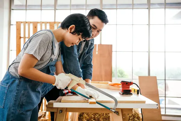 stock image Asian father and son work as a woodworker or carpenter, Father teaches his son to saw a wooden plank with hack saw carefully together with teamwork. Craftsman carpentry working at home workshop studio