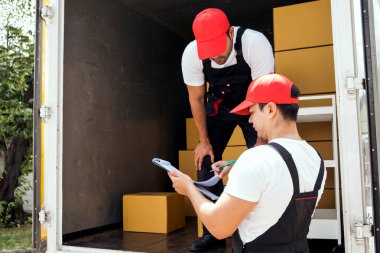 Üniformalı Asyalı ve Kafkasyalı işçiler kamyondan karton kutuları boşaltıyorlar. Teslimatçılar kutuları boşaltıyor ve iş arkadaşlarıyla kontrol ediyorlar. Profesyonel dağıtım ve taşıma hizmeti.