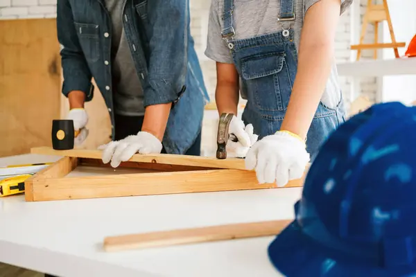 Stock image Two individuals work on a woodworking project, focused on crafting while wearing safety gear, with tools and materials spread out on a white table.