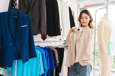 Bir kadın moda mağazasında giysi eşyasıyla poz veriyor, çeşitli renkli giysiler sergileniyor..