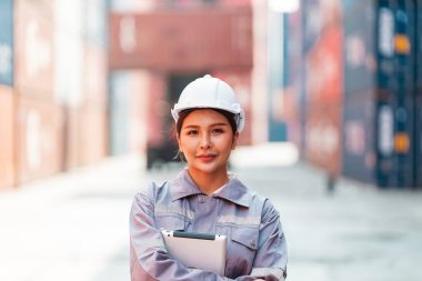 Kasketli ve iş üniformalı kendine güvenen Asyalı bir kadın bir tersanede elinde bir not panosuyla duruyor, profesyonelliği ve lojistikteki kararlılığı sembolize ediyor. Nakliye işletmesi yönetimi.