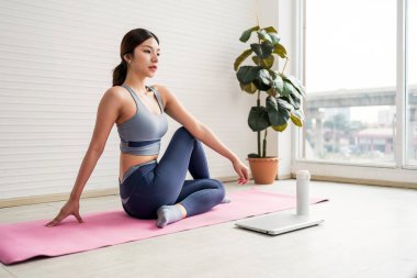 Bir kadın pembe bir paspasın üzerinde yoga yapıyor. Parlak, ferah bir odada huzurlu bir ifadeyle oturarak poz veriyor..