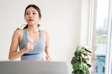 Genç bir kadın evde egzersiz yapıyor, pencerenin yanında koşuyor, arka planda bir dizüstü bilgisayar ve bitkiyle spor yapıyor..
