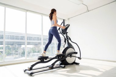 Parlak, modern bir spor salonunda eliptik bir makine üzerinde egzersiz yapan bir kadın, spor salonuna ve vücut sağlığına odaklanıyor..