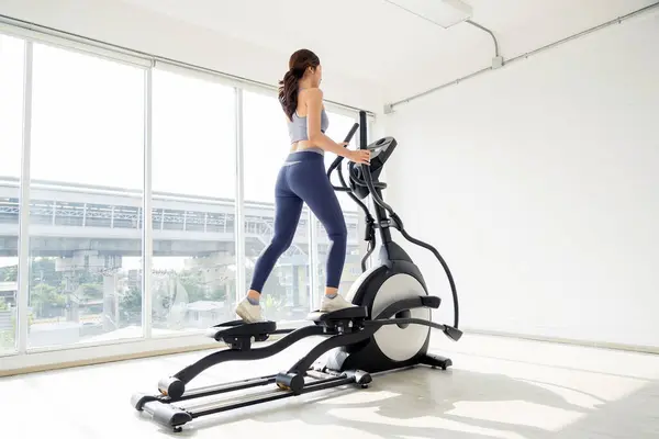 Parlak, modern bir spor salonunda eliptik bir makine üzerinde egzersiz yapan bir kadın, spor salonuna ve vücut sağlığına odaklanıyor..