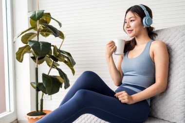 A woman in active-wear relaxes on a couch, sipping coffee and wearing headphones, with a potted plant nearby. clipart