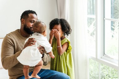 Neşeli bir baba iki küçük çocuğuyla etkileşime girer, biri kucağında oturur ve diğeri neşeyle gözlerini kapatır, sıcak bir aile anı yaratır. Mutlu çeşitlilik aile kavramı.