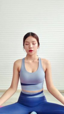 Huzurlu bir Asyalı kadın yoga yapıyor. Gözlerini kapalı bir alanda açıyor. Yoga minderinde rahatlık ve farkındalık simgeliyor. Evde çalışma konsepti. Yavaş çekim 4k video.