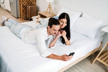 Bir çift, rahat bir şekilde bir yatakta uzanmış, akıllı bir telefona dalmış, rahat ve modern bir yatak odasında, bir anlık bir bağlantı ve neşe paylaşıyor..