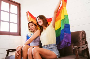 İki genç kadın sevinçle bir gökkuşağı bayrağı sergilerken bir kanepede oturmuş gururu ve kapsayıcılığı coşkulu ve neşeli bir ortamda kutluyorlar. LGBTQIA çiftler ilişkileri birlikte kaliteli zaman geçirirler..