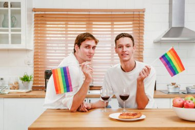 İki beyaz erkek, sevgi ve gururu kucaklayan sıcak bir mutfakta gökkuşağı bayrakları ve şarap kadehleriyle kutlama yapıyor. LGBTQIA çiftler ilişkileri birlikte kaliteli zaman geçirirler..
