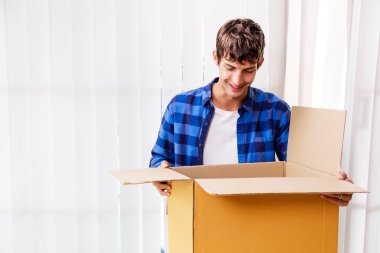 Genç bir adam içerideki bir karton kutuyu açarken gülümsüyor ve içindekileri gösteriyor. Mutlu müşteri karton kutu açmak Online mağaza siparişi ile, iyi nakliye hizmeti