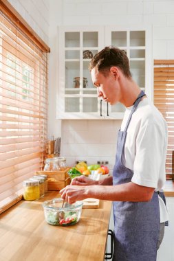 Genç bir adam renkli meyve ve sebzelerle çevrili parlak bir mutfakta sağlıklı bir yemek pişirme tarzı sergileyen taze bir salata hazırlıyor. LGBT ilişkileri. Eşcinsel çift yemek pişirme konsepti.