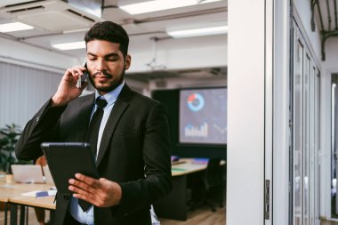Takım elbiseli profesyonel bir adam, modern bir ofis ortamında tableti kontrol ederken bir telefon görüşmesi yapar. Toplantıda Asyalı işadamı rolü.