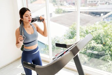 Asyalı genç bir kadın, elinde su şişesiyle koşu bandında egzersiz yapıyor ve antrenmanına odaklanıyor. Spor kıyafetleri giyerek evde spor yapıyor ya da egzersiz yapıyor..