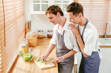 Happy caucasian gay couple making salad together One person used a knife to cut lettuce in front. And another person watching from behind in the kitchen at home. LGBT relationships. Gay couple concept