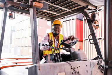 Kaskı ve güvenlik yeleği giyen gülümseyen Afrikalı bir forklift operatörü nakliye konteynırlarıyla dolu bir açık hava deposunda onay veriyor. Konteynırları dikkatlice taşımaya odaklanmış.