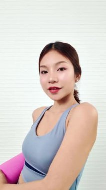 Genç bir kadın, spor kıyafeti giymiş pembe bir yoga minderi tutuyor. İyi aydınlatılmış bir yerde antrenmana hazır. Spor kıyafetleri giyerek evde spor yapıyor ya da egzersiz yapıyor..