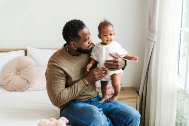 Sevgi dolu bir baba, bebeğini sıcak ve şefkatli bir anı paylaşan parlak bir pencerenin yanında tutar. Sıcak aile sahnesi, güven ve sevgiyle dolu evde babalığı, yakınlığı, ilgiyi ve sevgiyi yakalar..